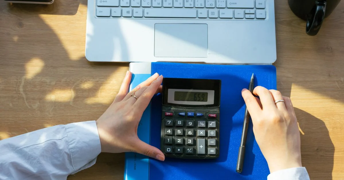 Manos usando una calculadora en un escritorio con un portátil, pluma y cuaderno. Perfecto para temas de oficina o finanzas.