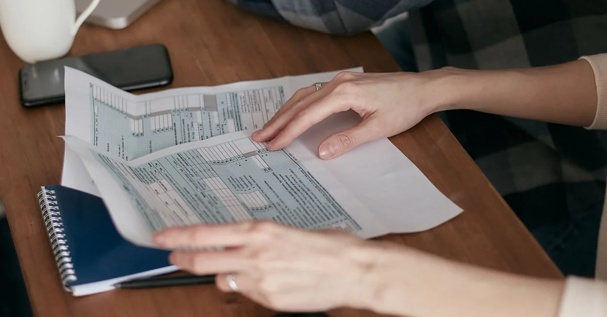 Acercamiento de manos organizando formularios de impuestos en un escritorio con una calculadora, portátil y cuaderno.
