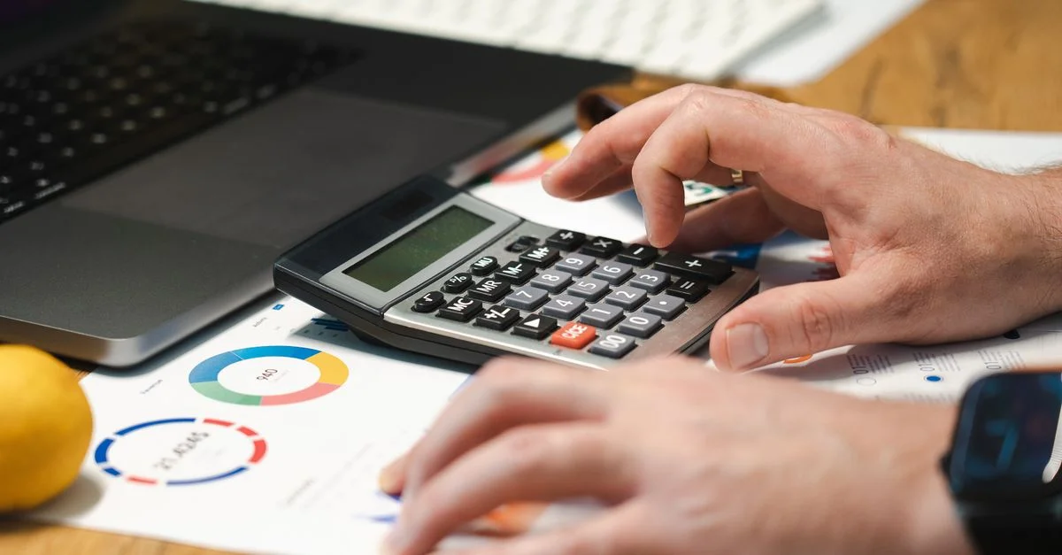 Una persona analizando datos financieros con una calculadora y una laptop en un escritorio.