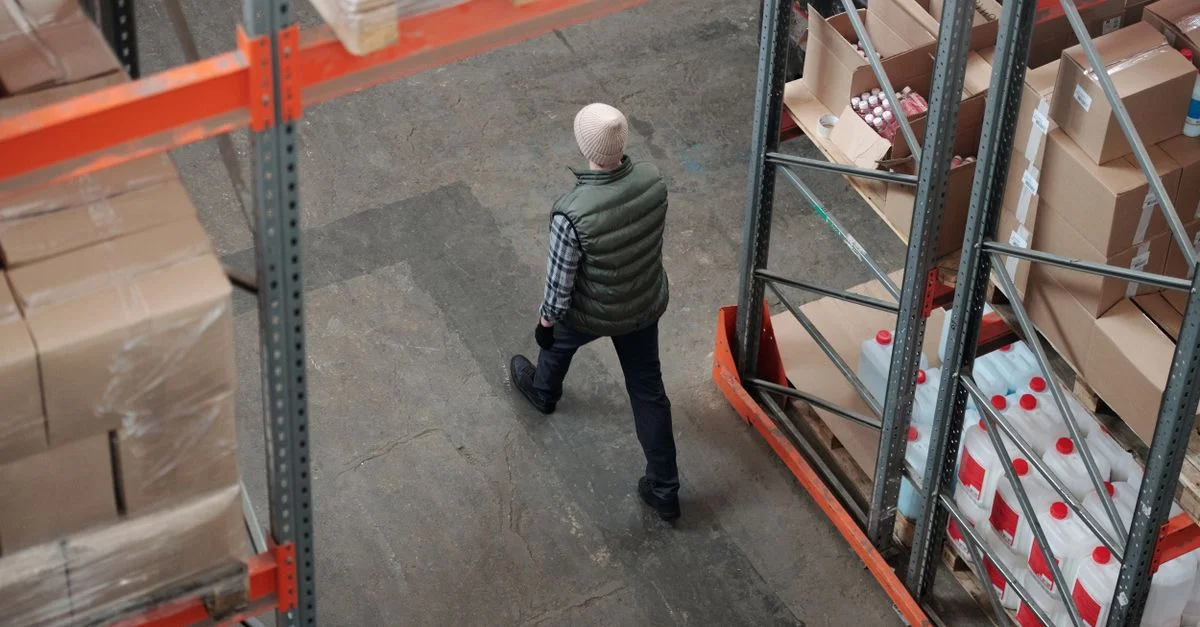 Vista aérea de un trabajador caminando por un pasillo de almacén rodeado de estanterías de paquetes.