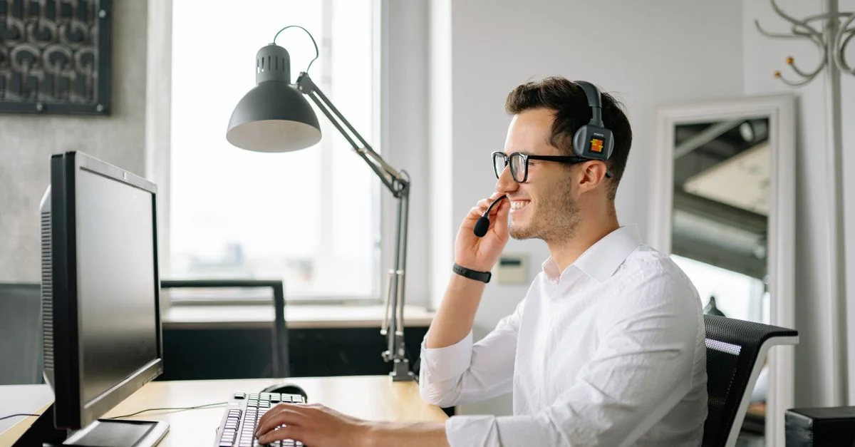Hombre sonriente usando auriculares trabajando en la computadora en una oficina luminosa.